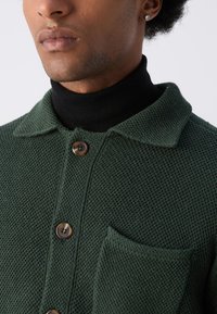 Cárdigan verde de punto con un acabado texturizado, presenta un collar, tres botones marrones y un bolsillo frontal. Lleva puesto sobre un cuello de tortuga negro.