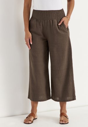 Kvinde iført brune vidde cropped bukser med elastisk smocktalje, hvid top og brune åbne sandaler, stående med den ene hånd i lommen.