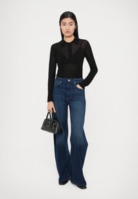 Svart langermet topp med krage, kombinert med vide blå jeans. Modellen holder en liten svart håndveske. Enkel, moderne design.