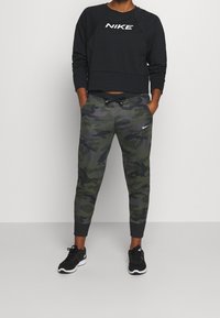 Musta lyhyt collegepaita, jossa on valkoinen "NIKE"-logo, yhdistetty vihreisiin maastokuvioisiin collegehousuihin, joissa on kiristysnyöri vyötäröllä, kapenevat lahkeet ja mustat lenkkarit.