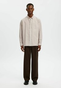 Ανοιχτό μπεζ πουκάμισο με κουμπιά, τσέπη στο στήθος, μακριά μανίκια και άνετη εφαρμογή, σε συνδυασμό με καφέ παντελόνι με φαρδιά πόδια και μαύρα παπούτσια.