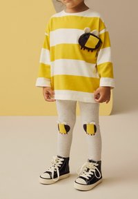 Gelb-weiß gestreiftes Langarmhemd mit einer Tasche, die ein Käferdesign zeigt; kombiniert mit grauen Leggings mit Käfer-Patches und schwarzen High-Top-Sneakers.