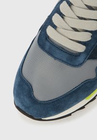 Σκούρο μπλε σουέντ αθλητικό παπούτσι με γκρι �πάνελ από δίχτυ, με επίπεδα μπεζ κορδόνια, στρογγυλή μύτη και νεονέ κίτρινη λεπτομέρεια στη σόλα.