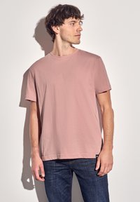 Camiseta de manga corta color rosa claro, cuello redondo, corte recto, fabricada en tejido suave, combinada con jeans azul oscuro, con un branding mínimo en el dobladillo.