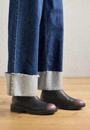 Zbliżenie na czarne skórzane botki noszone z szerokimi jeansami podwiniętymi, aby odsłonić jasne mankiety z denimu na drewnianej podłodze.