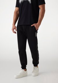 Schwarze Jogginghose mit schlanker Passform, weißen Seitenstreifen und sichtbar logo "GUESS" auf dem linken Oberschenkel. Kombiniert mit weißen Turnschuhen.