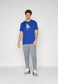Kék póló fehér "LA" logóval és Nike pipa emblémával, szürke nadrággal és fekete sportcipővel, egyszínű háttér előtt állva.