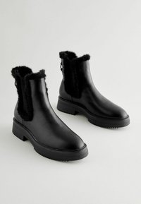 Botas negras hasta el tobillo hechas de cuero liso con forro de pelaje. Diseño elegante, punta redondeada y suela de goma texturizada para mayor agarre.