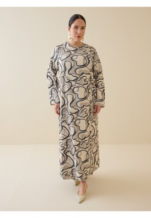Kvinde iført en lang beige kjole med sorte abstrakte svirvler, lange ærmer og beige spidse hæle på en ensartet lys baggrund.
