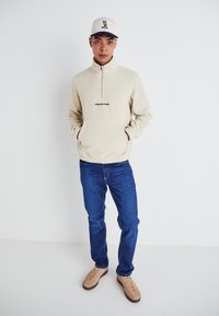 Beige fleece quarter-zip pullover with front pocket, featuring "Original Studio" text. Paired with blue jeans and beige sneakers, on a neutral backdrop.
