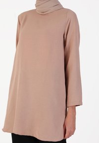 Lys beige tunika med høj hals og lange ærmer, lavet af blødt, tekstureret stof. Har en forlænget kant og en løs pasform.
