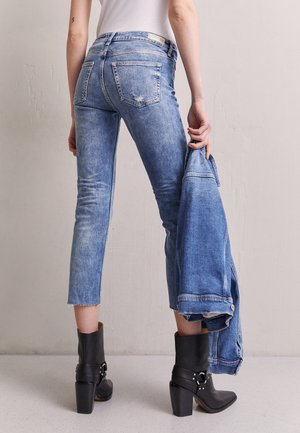 Kobieta ubrana w krótkie niebieskie dżinsy i czarne botki na obcasie, trzymająca w ręku dopasowaną niebieską kurtkę dżinsową na tle gładkiej ściany.