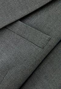Nahaufnahme einer grauen Anzugjackentasche mit strukturiertem Stoff und sichtbaren Nähdetails.