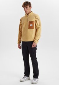 Geel fleece trui met halve ritssluiting, bruine borstzak en zijritszak. Zwarte broek en witte sneakers maken de outfit compleet.