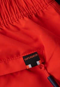 Pantalones cortos rojos brillantes con cintura fruncida y detalle de cordón; presenta una etiqueta negra con "SUPERDRY" en acentos naranja y blanco.