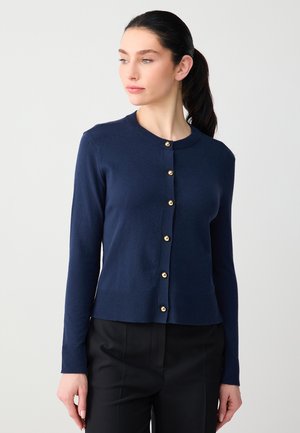 Donna con capelli scuri raccolti in una coda di cavallo, indossa un cardigan blu navy con bottoni dorati e pantaloni neri, guarda verso sinistra su uno sfondo neutro.