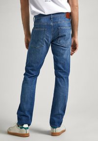 Vaqueros de mezclilla azul con corte recto, que cuentan con doble bolsillo trasero y una tapeta de cuero marrón en la cintura.