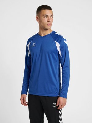 Ung mand iført en blå langærmet sportstrøje og sorte bukser med hvide chevron-mønstre, står og kigger til siden.