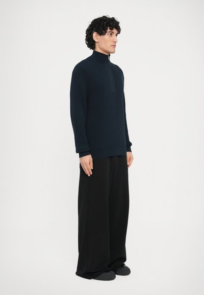 Pull-over côtelé bleu marine avec un col haut et une fermeture éclair à moitié. Porté avec un pantalon noir ample et des chaussures foncées. Look simple et moderne.