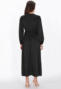 Schwarzes, langärmeliges Kleid mit geraffter Taille. Glatter, satinähnlicher Stoff, knöchellang, mit hohem Ausschnitt und fließender Silhouette.