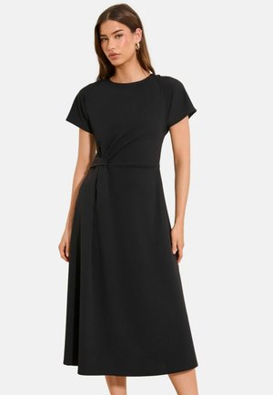 Žena v čiernych midi šatách s krátkym rukávom a detailom uzla na boku, stojaca pred jednoliatym bielym pozadím.
