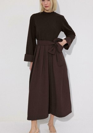 Mujer con un vestido midi de manga larga color marrón oscuro, falda plisada, puños anchos y un gran lazo atado en la cintura, de pie contra una pared blanca.