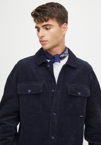 Navy corduroy shirt jacket with a button-up front, two chest pockets, and a ribbed texture. Worn over a white shirt with a blue bandana.