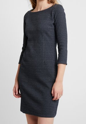 Frau trägt ein tailliertes dunkelblau gemustertes Kleid mit Dreiviertelärmeln und steht vor einem einfarbigen hellen Hintergrund.