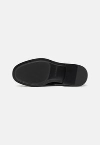 Suela de zapato sintética negra con textura suave, forma redondeada y detalles de tracción. Presenta un ligero tacón y una marca mínima.