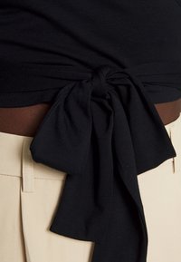 Černý látkový top s detailem zavázané uzlu v pase, doplněný světle béžovými kalhotami s vysokým pasem, které mají viditelné švy a kapsy.