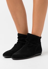 Černé kotníkové boty vyrobené z měkkého semiše, s uvolněným designem, zaoblenou špičkou a nízkou, odolnou podrážkou. Hladký povrch, žádné viditelné kování.