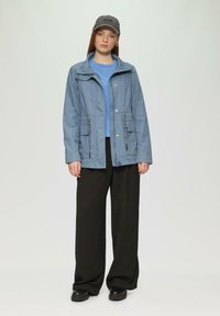 Giacca blu chiaro con zip e due tasche frontali, abbinata a una camicia blu e pantaloni neri a gamba larga. Cappellino nero e scarpe nere chunky.