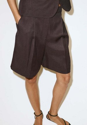 Persona con pantalones cortos a medida de color marrón oscuro hasta la rodilla y sandalias negras con tiras en el tobillo, de pie con las manos en los bolsillos frente a un fondo liso.