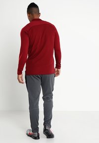 T-shirt de sport rouge à manches longues et ajustée avec des détails sur les panneaux latéraux, associé à un pantalon élastique gris et des chaussures de sport noires et rouges.