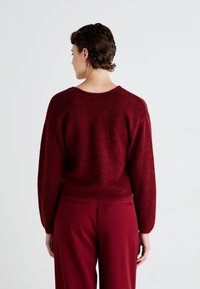 Rust-colored, soft knit sweater with a wide neckline and dropped shoulders, worn with high-waisted, matching red pants.