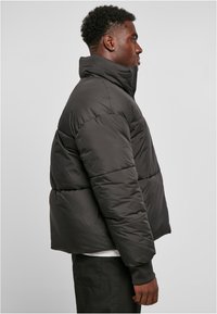 Chaqueta de plumas negra con un alto cuello, acolchado horizontal y puños elásticos, diseñada con una silueta corta para un ajuste ceñido.