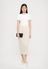 Haut blanc à manches courtes associé à une jupe beige texturée allant jusqu'aux genoux. Tenant une pochette noire et portant des chaussures blanches à bout pointu.