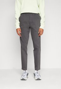 Grijze, lichte broek met een elastische tailleband, taps toelopende pijpen en een gladde textuur. Gecombineerd met witte en grijze sportschoenen.
