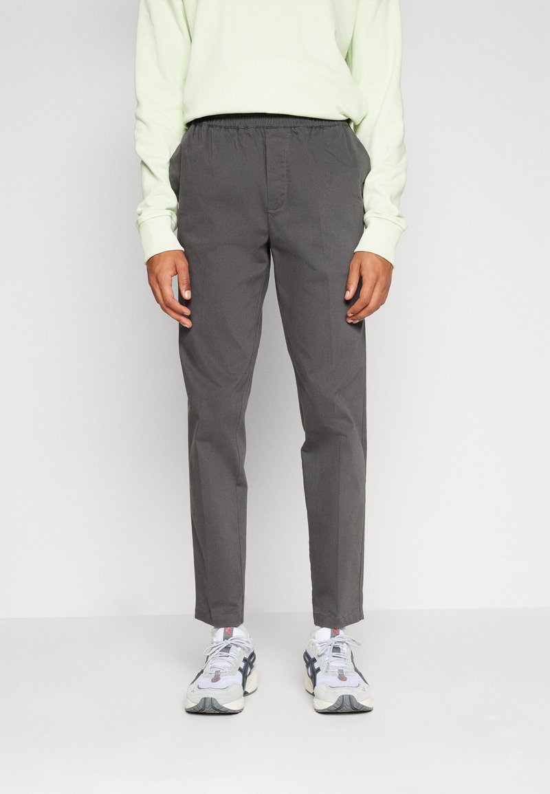 Grijze, lichte broek met een elastische tailleband, taps toelopende pijpen en een gladde textuur. Gecombineerd met witte en grijze sportschoenen.