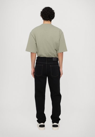 Camiseta de gran tamaño verde oliva con mangas cortas, combinada con vaqueros rectos negros que presentan un diseño clásico de cinco bolsillos y costuras en tono marrón claro.