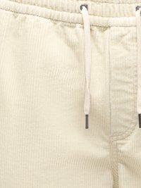 Gros plan d’un pantalon en velours côtelé crème avec une taille élastique et des cordons blancs avec embouts métalliques.