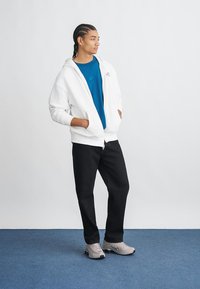 Biela mikina na zips s kloktami, prehodená cez modré tričko. Čierne nohavice a svetlošedé tenisky dopĺňajú celý outfit.