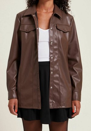 Vrouw draagt een bruine imitatieleren jas met knopen over een wit geribbeld shirt, zwarte rok en visnetkousen, staand met ontspannen handen.