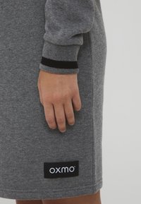 Siva haljina od dukserice s rebrastim crnim detaljima na manžetama, sa logotipom oxmo na crnoj oznaci blizu poruba. Mekani tekstil.