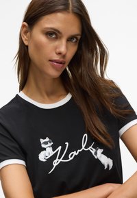 Mulher com cabelo castanho longo vestindo uma t-shirt preta com um desenho branco e dois gatos brancos de óculos escuros ao redor do texto em caligrafia "Karl".
