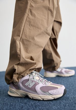 Person iført beige bukser og lilla New Balance-sneakers, der står på blåt tæppe.