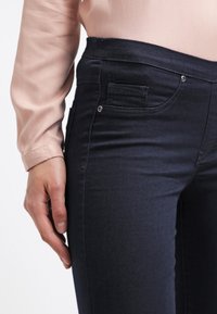 Tummansinisiä farkkuja, joissa on sileä pinta, pienellä etutaskulla ja puolikorkealla vyötäröllä, yhdistettynä vaaleanpunaiseen puseroon.