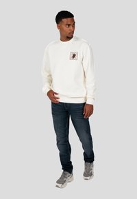 Felpa crema chiaro con un patch quadrato stampato sul petto, abbinata a jeans blu scuro e sneakers grigie. Il design è semplice e classico.