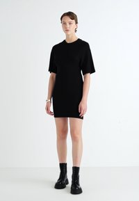 Melnas īsām piedurknēm kleita ar pieguļošu siluetu, izgatavota no mīkstas auduma, apvienota ar melniem šņorējamiem zābakiem. Minimālistisks dizains un tekstūra.