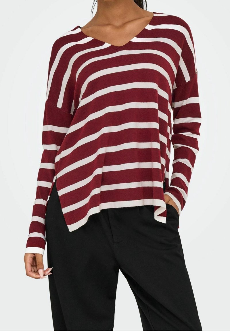 Femme portant un haut ample à manches longues rayé bordeaux et blanc avec un pantalon noir, une main dans la poche et l'autre détendue le long du corps.